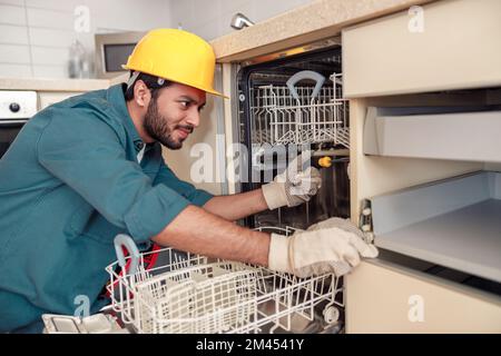 Focused handyman in special clothing repairing dishwasher in modern kitchen Stock Photo