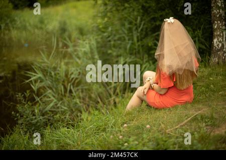 Girl on shore of lake in summer. Maiden in orange clothes. Bride in dress. Holiday in park. Beautiful clothes. Stock Photo