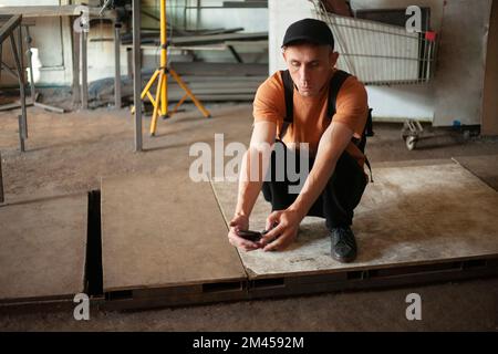 Guy is sitting on floor in garage. Guy looks at phone. Man is waiting for work. Man is squatting. Stock Photo