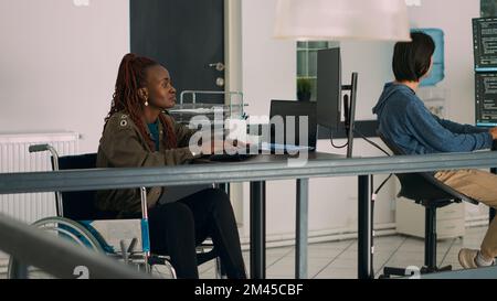 Cloud programmer in wheelchair using server data to create software app on computer, suffering from chronic disability. Coder working on new use rinterface development in agency office. Stock Photo