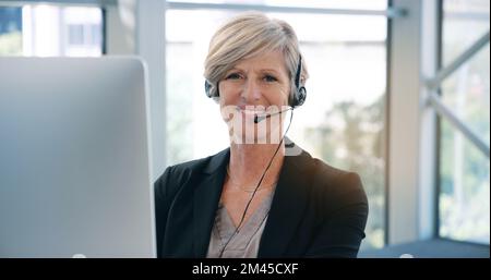We take the time to chat to you. Portrait of a mature businesswoman wearing headphones while working on a computer in an office. Stock Photo