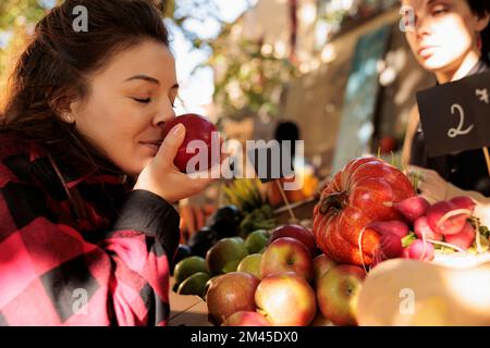 Healthy customer enjoying organic natural smell of apples, standing in front of farmers market stand. Woman smelling bio fruits before buying homegrown eco produce from counter. Stock Photo