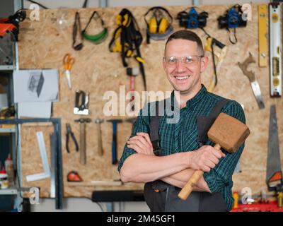 Portrait of a carpenter in goggles and overalls holding a wooden hammer in the workshop against the background of a wall with tools. Stock Photo