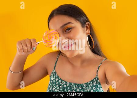 Closeup portrait of a teenage girl holding lollipop over her eye against yellow background Stock Photo