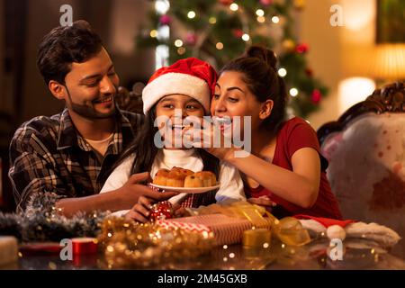 Mother feeding cupcakes to her daughter on the occasion of Christmas while father holding plate Stock Photo
