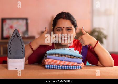 Close-up portrait of a happy woman showing thumbs up next to ironed shirts Stock Photo