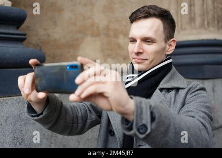 Handsome young man in coat walking on street holds mobile phone and shoots video or takes photo, takes a selfie on smartphone and communicates online via video communication Stock Photo