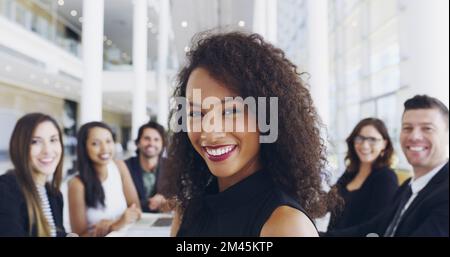 I earned my place at the head of the table. a young businesswoman smiling in an office during a meeting with her colleagues in the background. Stock Photo
