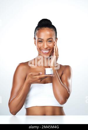This is the best stuff. Studio portrait of an beautiful young woman applying skin moisturizer to her face while standing next to a white background. Stock Photo