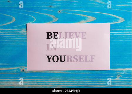 Believe in yourself sign fading into a Be you message written on pink paper placed over textured blue wooden background. Stock Photo