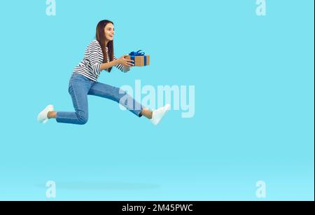 Happy joyful woman holding gift box, jumping and flying on blue copy space background Stock Photo