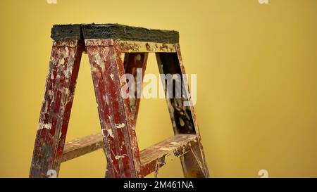 Old used construction painter ladder with painted splatter standing in front of the yellow wall under warm white lighting Stock Photo