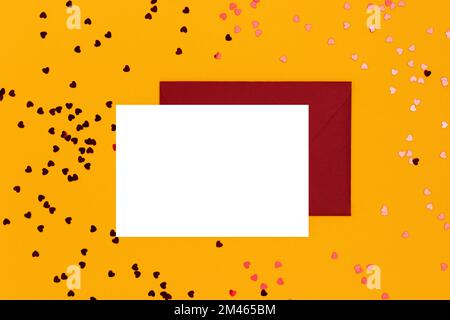 Empty white paper mockup, envelope and red hearts confetti on a yellow background. Creative concept. Stock Photo