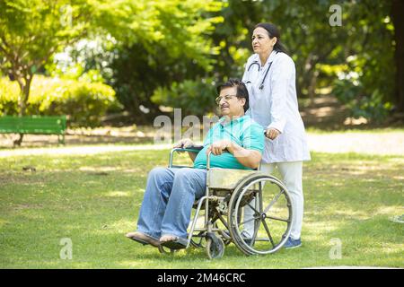 Indian caregiver nurse taking care of senior male patient in a wheelchair outdoor at park, Asian doctor help and support elderly mature older people. Stock Photo