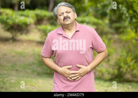 Indian senior man having stomachache or diarrhea outdoor at park, Asian Old male suffering from abdominal pain, health problem. Stock Photo