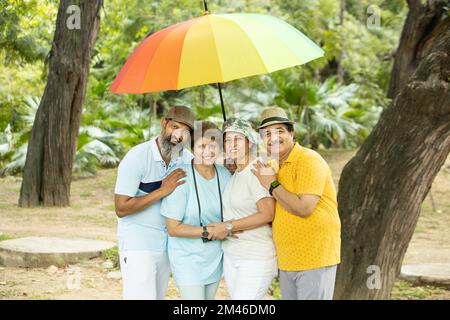 Group of happy indian senior people standing under big colorful umbrella wearing casual cloths and hat outdoor at park, Retired old people enjoying ho Stock Photo