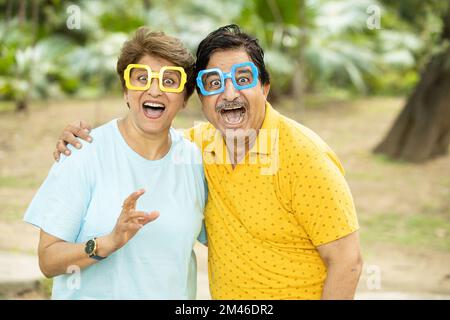 Portrait of crazy excited funny senior indian couple wearing funky eye glasses outdoor at park. Mature people making faces. Stock Photo