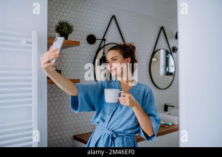 Young woman taking selfie in bathroom, enjoying cup of coffee. Stock Photo