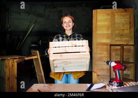 Woman holding wooden crate in her hands and smiling while working in carpentry workshop. Stock Photo