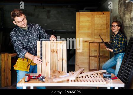 Woman using laptop spending time with her husband while he working in his workshop. Stock Photo