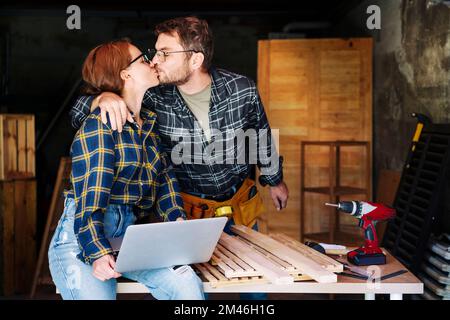 Young adult couple kissing celebrating success of their small business while working in their carpentry workshop. Stock Photo