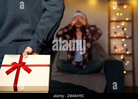 Christmas present boxes in hand behind back defocused woman with closed eyes sitting on sofa and waiting xmas gifts. Stock Photo