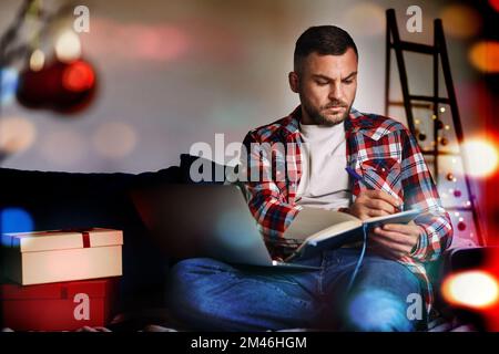 Young man working alone at home while Christmas night using laptop and writing notes in his notebook. Stock Photo