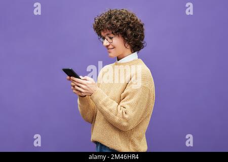 Side view of happy young brunette woman in eyeglasses and beige pullover using smartphone while standing on violet background Stock Photo