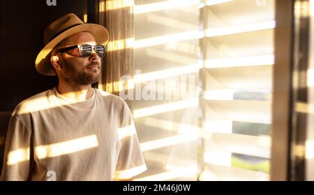 Casual clothing man with a short beard wearing a straw hat and sunglasses standing in the room and looking through the window blinds. Stock Photo