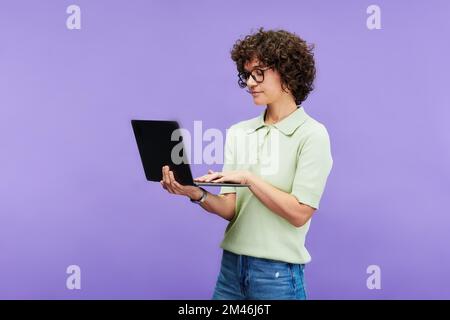 Young serious brunette woman in casualwear typing on laptop keyboard while networking on studying online against lilac background Stock Photo