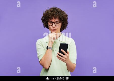 Young pensive woman with smartphone looking through goods in online shop and trying to make choice between some of them Stock Photo
