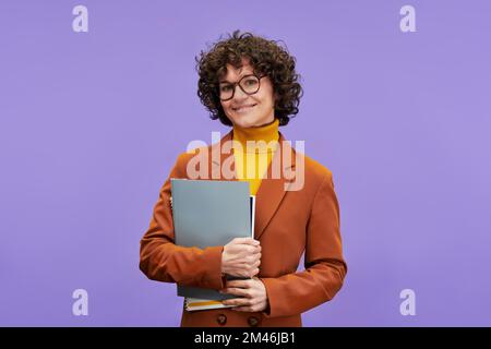 Young successful teacher in formalwear holding documents and looking at camera while standing on lilac background in isolation Stock Photo