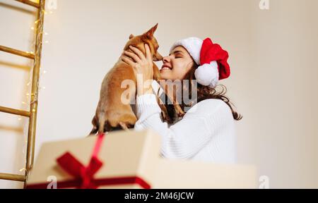 Happy woman wearing Santa hat holding small dog in her hands Christmas gift. Stock Photo