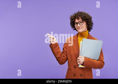 Young smiling woman with short curly brown hair looking at camera while standing against violet background and pointing at copyspace Stock Photo
