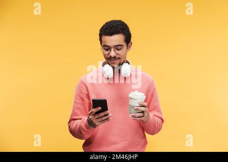 Young multi-ethnic man with white headphones around neck using smartphone and having cup of coffee against yellow background Stock Photo