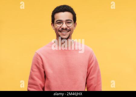 Young cheerful man in eyeglasses and pink sweatshirt looking at camera while standing against vivid yellow background in isolation Stock Photo