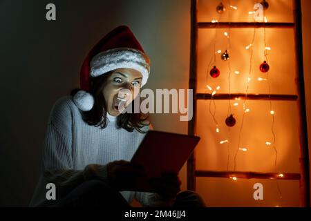 Happy woman in Santa hat holding digital tablet in her hands looking at screen and screaming with happiness and surprise. Stock Photo