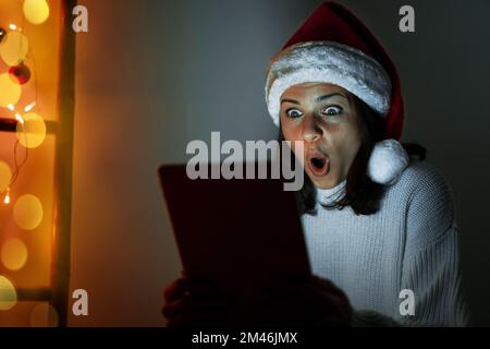 Woman in Santa hat holding digital tablet in her hands and looking at screen in surprise with wide eyes. Stock Photo