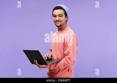 Young successful male freelancer or programmer in pink pullover, beanie hat and eyeglasses holding laptop and looking at camera Stock Photo