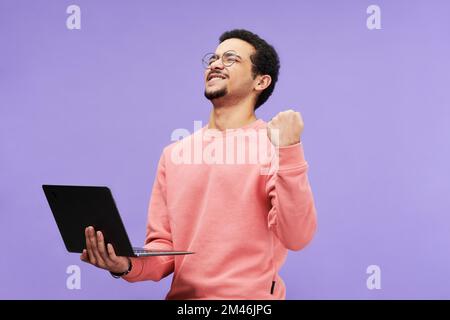 Ecstatic guy in pink casualwear keeping eyes closed while holding laptop and expressing excitement against violet background in isolation Stock Photo