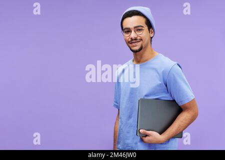 Young smiling businessman or student in blue t-shirt and beanie holding laptop while standing on violet background and looking at camera Stock Photo