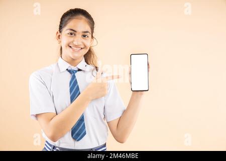 Happy Indian student schoolgirl wearing school uniform holding smart phone and pointing, blank screen to put advertisement isolated over beige backgro Stock Photo