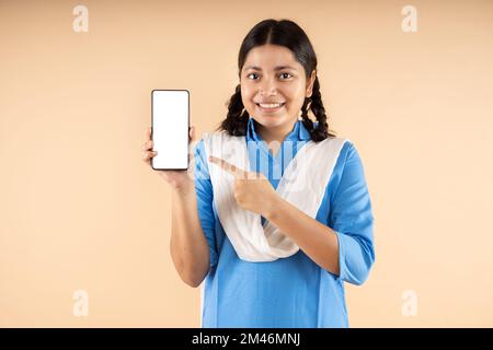 Happy rural Indian student schoolgirl wearing blue government school uniform holding smart phone and pointing, blank screen isolated on beige backgrou Stock Photo
