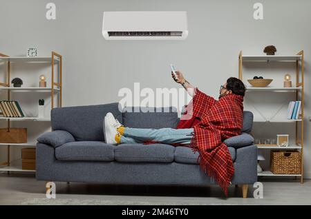 Young man wrapped in blanket holding air conditioner remote control Stock Photo