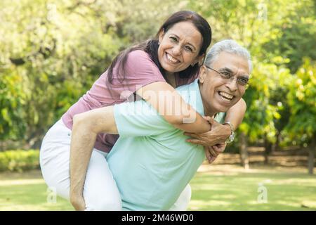 Happy indian senior couple enjoying life having fun at summer park, man giving piggyback ride to woman. Stock Photo