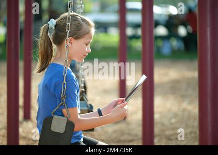 Happy child girl using digital tablet to play video game during summer vacations sitting on swing in park Stock Photo