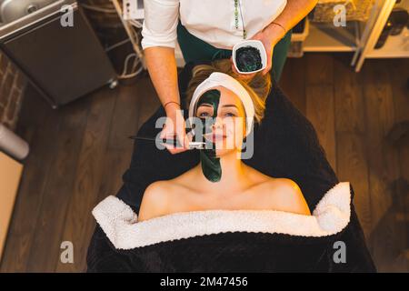 Happy SPA client on SPA bed under a blanket with charcoal mask. Unrecognizable beautician applying black mask with a brush. High angle indoor shot. Copy space. High quality photo Stock Photo