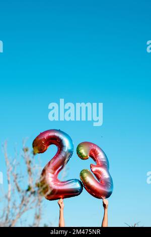 a man holds two multicolored number-shaped balloons forming the number 23 against the blue sky, with some blank space on top Stock Photo