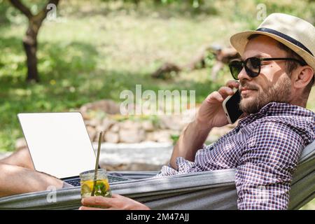 Freelancer man wearing hat and sunglasses calling phone, using laptop, drinking lemonade while lying in hammock. Stock Photo