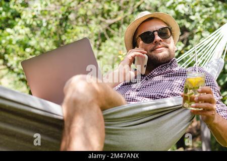 Freelancer using his phone and laptop lying in hammock, man in hat and sunglasses relaxing in hammock with drink and using digital devices. Stock Photo